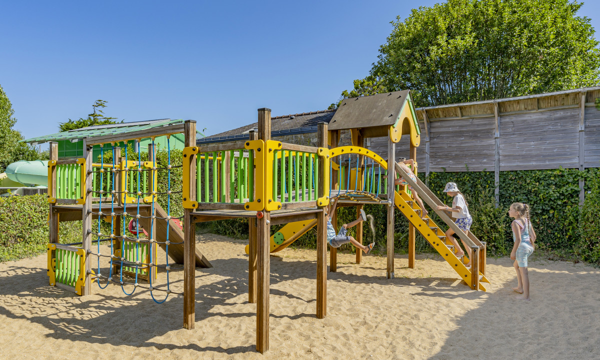 At De la Plage, kids enjoy themselves on a vibrant play structure featuring slides and climbing ropes, encircled by lush greenery and a wooden barrier.