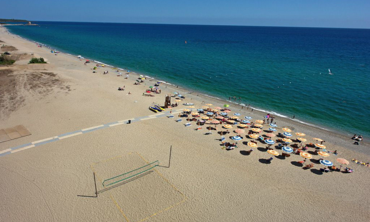 At Ultima Spiaggia, beach canopies shelter sunbathers close to the water's edge; a volleyball net remains unoccupied; the clear, azure sea extends outward.