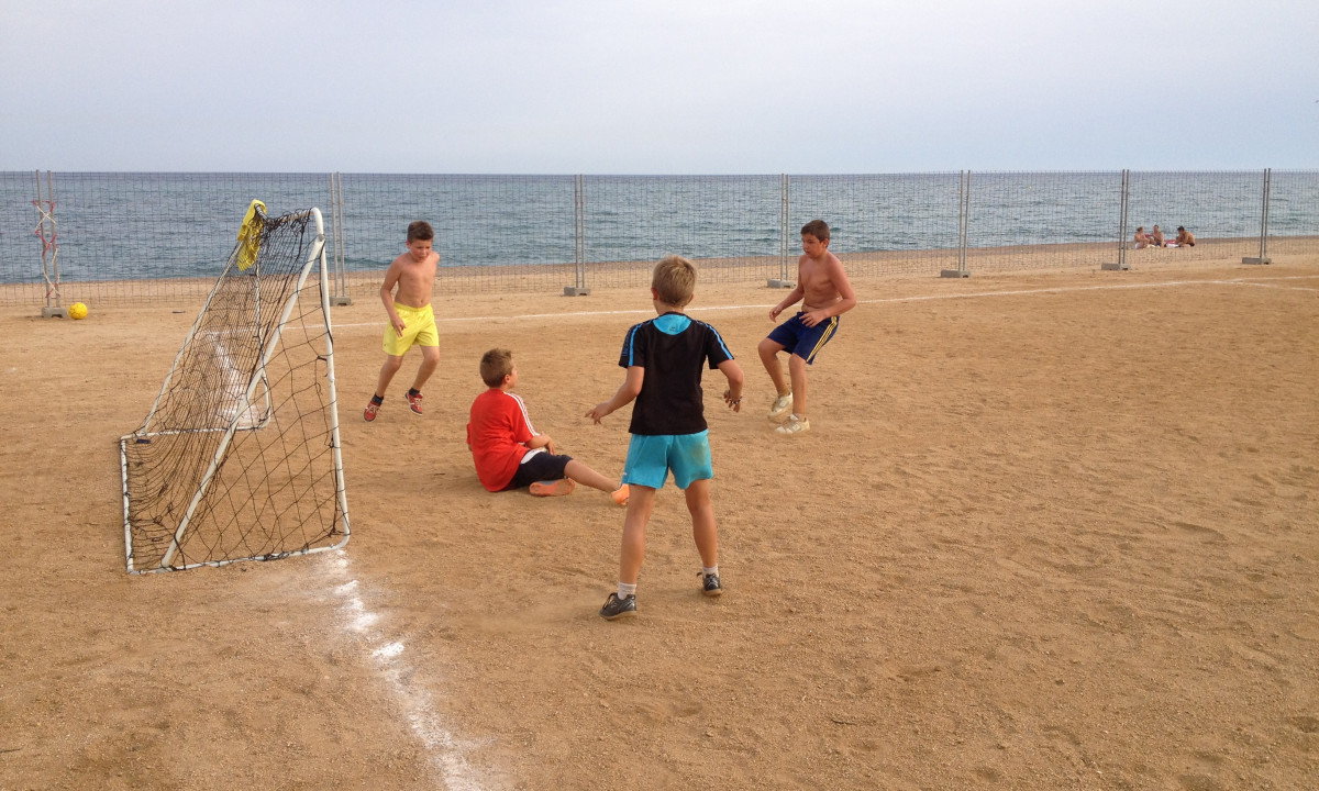 Four boys play soccer on a sandy field near a beach, with a goalpost and ocean in the background at Resort Els Pins
