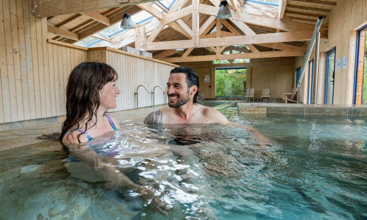 Two people relax and smile in an indoor pool, surrounded by wooden walls and a beamed ceiling at Maguide