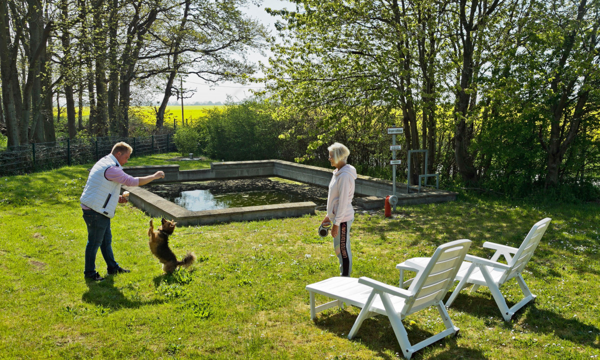 At KNAUS Campingpark Rügen, an individual interacts with a dog while another person watches nearby a rectangular pond encircled by trees and a grassy area.