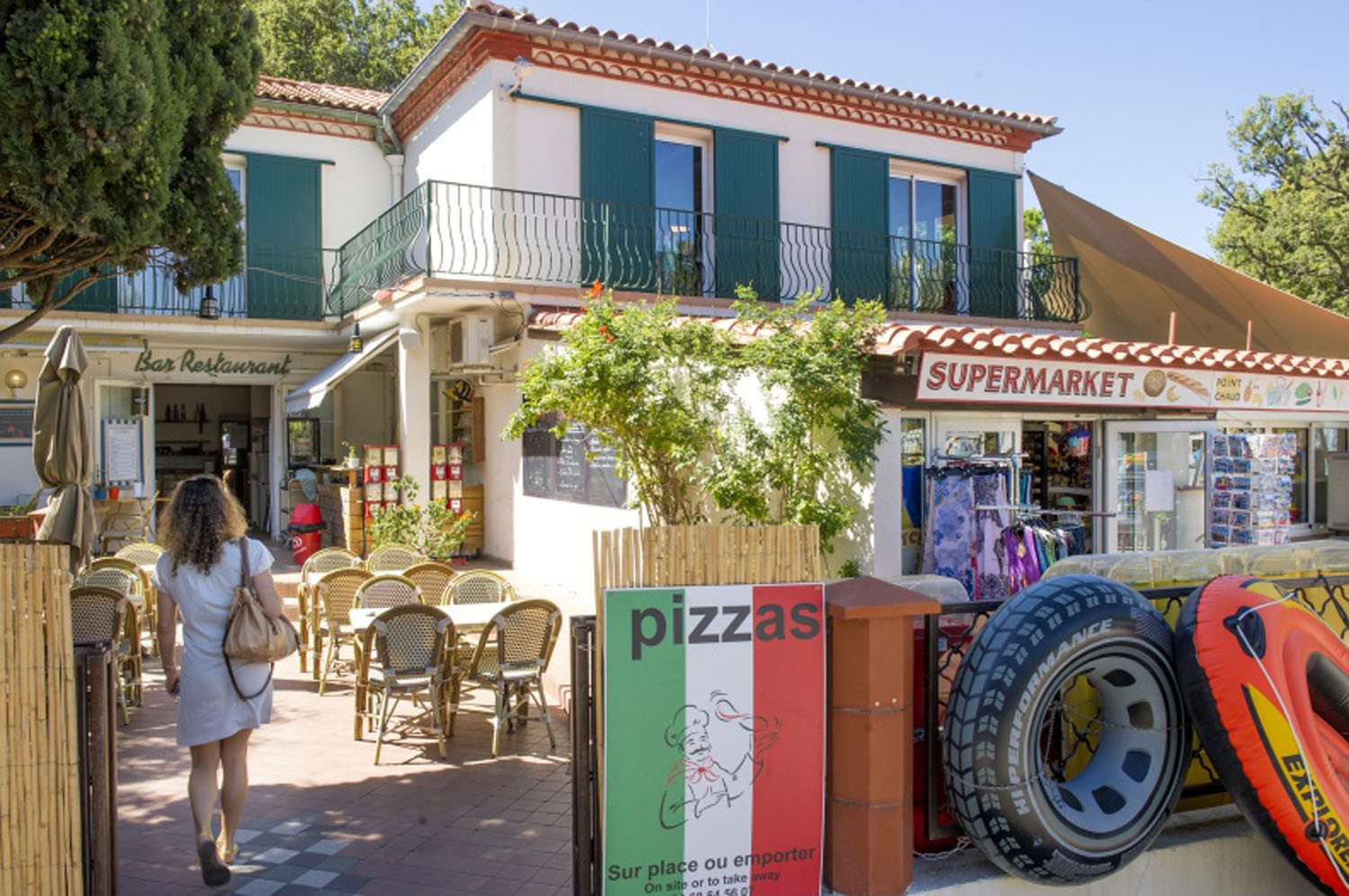 At Bois de Valmarie, a woman strolls toward the al fresco dining section outside a restaurant, with a grocery store to her right and tire tubes showcased nearby.