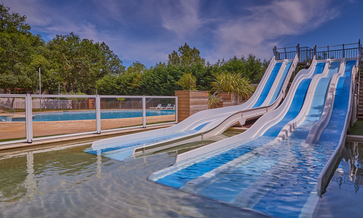 At Le Chêne Vert, azure and ivory water slides descend into a shallow basin, encircled by trees beneath a partly overcast sky.