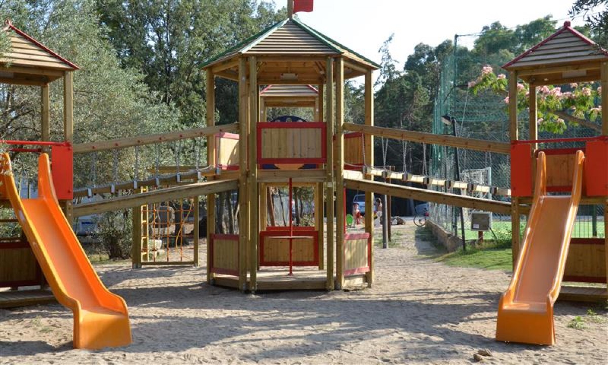 At Ultima Spiaggia, there is a wooden play structure featuring slides and bridges situated in a sandy play zone surrounded by trees and nearby individuals.