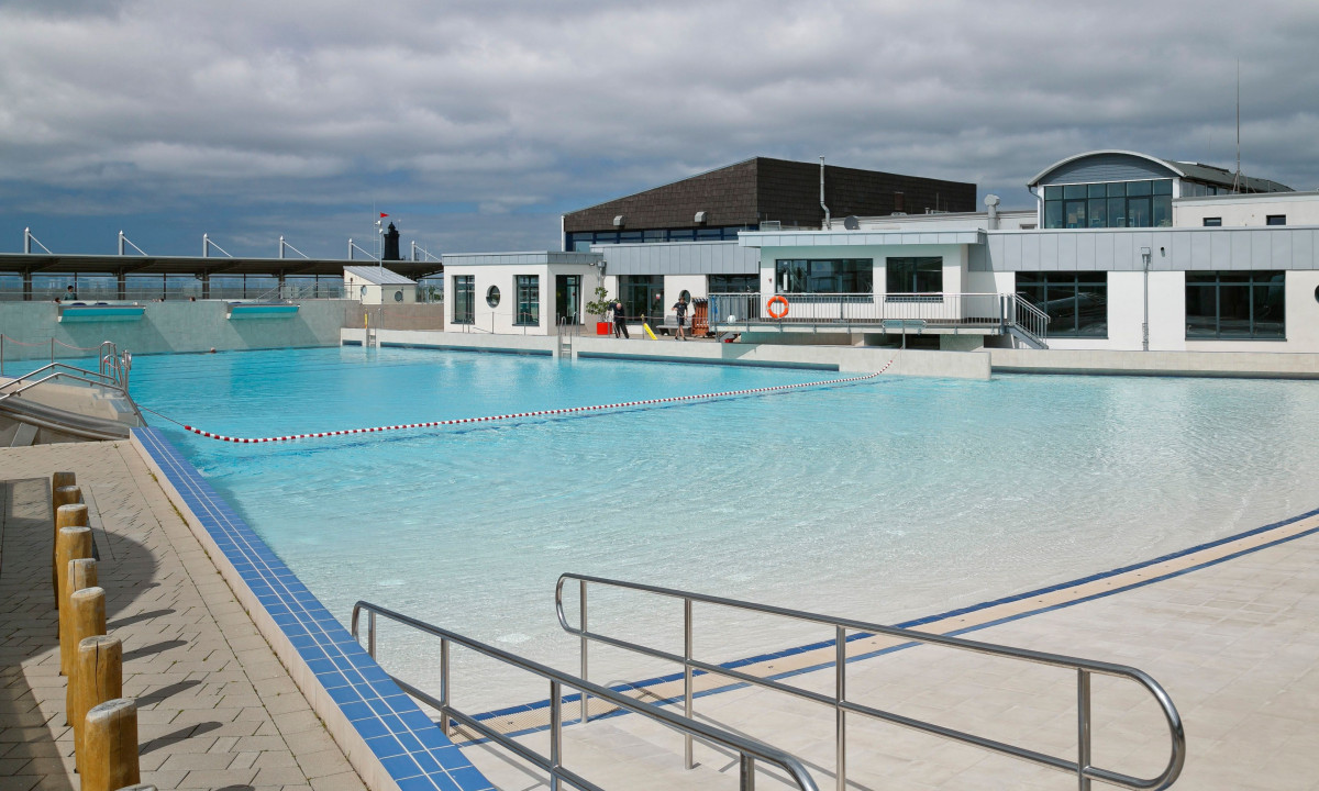 At KNAUS Campingpark Dorum, a spacious outdoor swimming pool with transparent water is surrounded by structures and walkways, all beneath a cloudy sky.