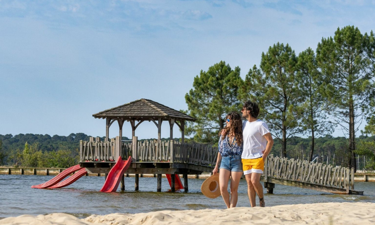 A couple strolls on a sandy lake beach near a wooden pavilion with slides; trees surround the area at Maguide