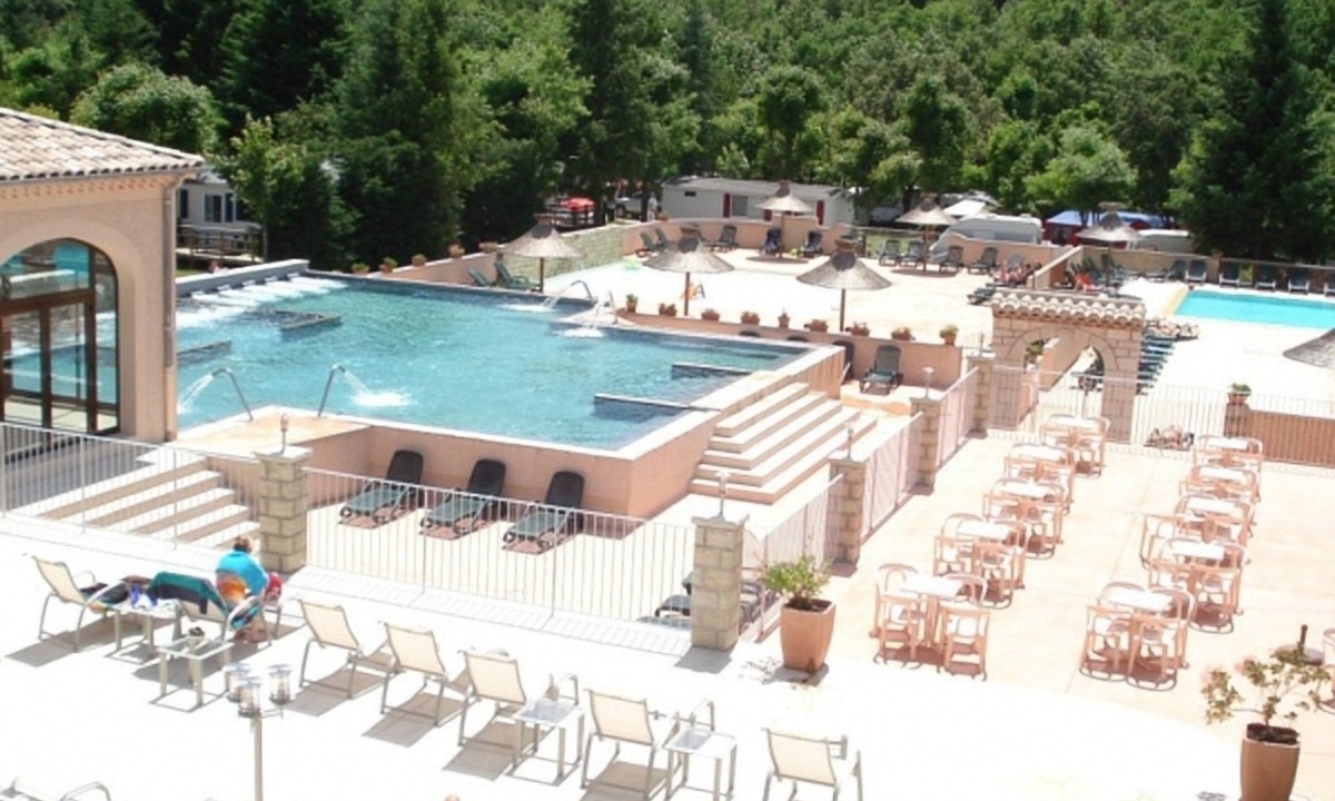 At Les Ranchisses, a swimming pool features lounge chairs and sun umbrellas in a resort-style environment surrounded by a vibrant, green forest.