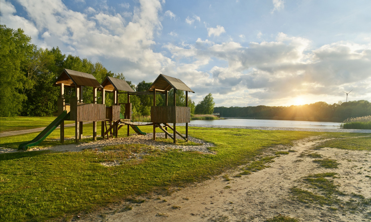 At KNAUS Campingpark Oyten, a wooden play structure remains idle by the lakeshore at dusk, encircled by greenery and trees.