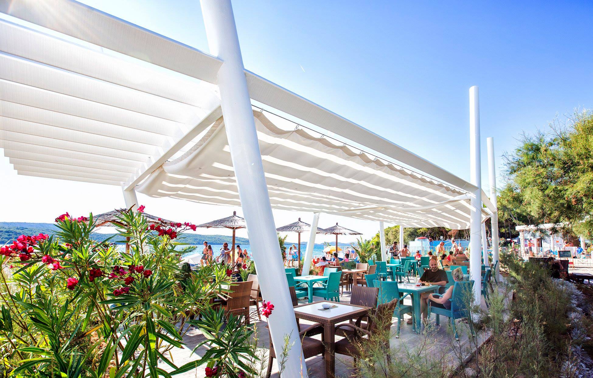 At Amadria Park Camping Sibenik, individuals are unwinding and enjoying meals beneath a spacious canopy, surrounded by beach umbrellas and vibrant seating, close to the ocean.