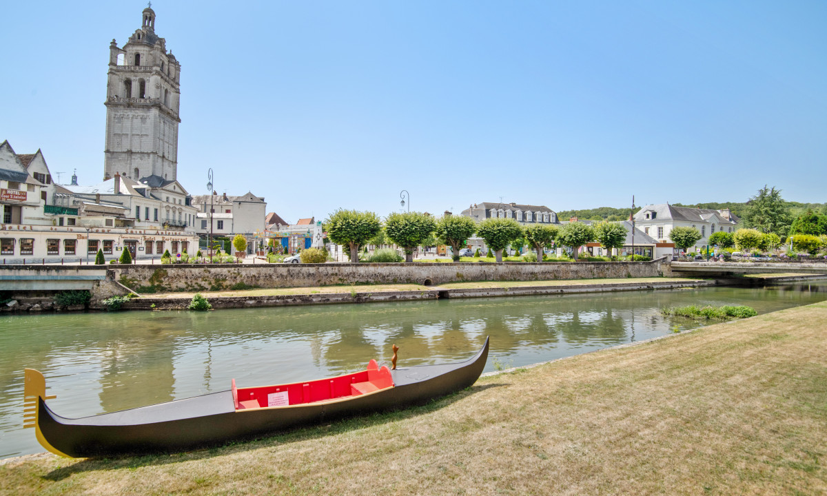 A gondola sits on a grassy riverbank, with a historic tower and town buildings across the water at Le Moulin des Cordeliers
