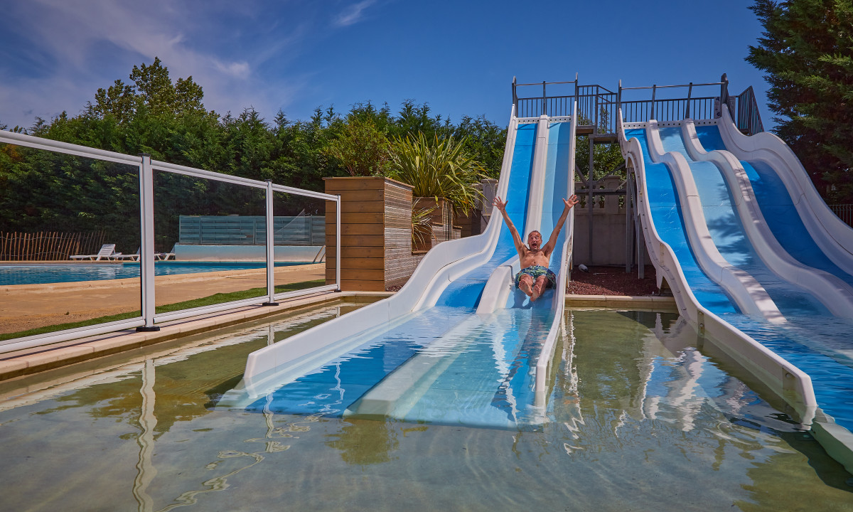 At Le Chêne Vert, an individual is gliding down a water slide with arms raised, surrounded by other slides and trees in the water park.
