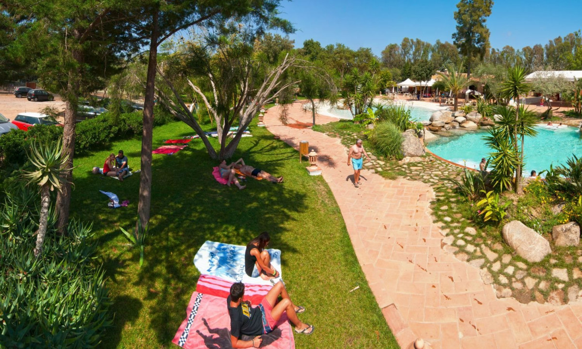 At Ultima Spiaggia, individuals are unwinding on towels and strolling along a pathway close to a swimming pool in a verdant, wooded region.