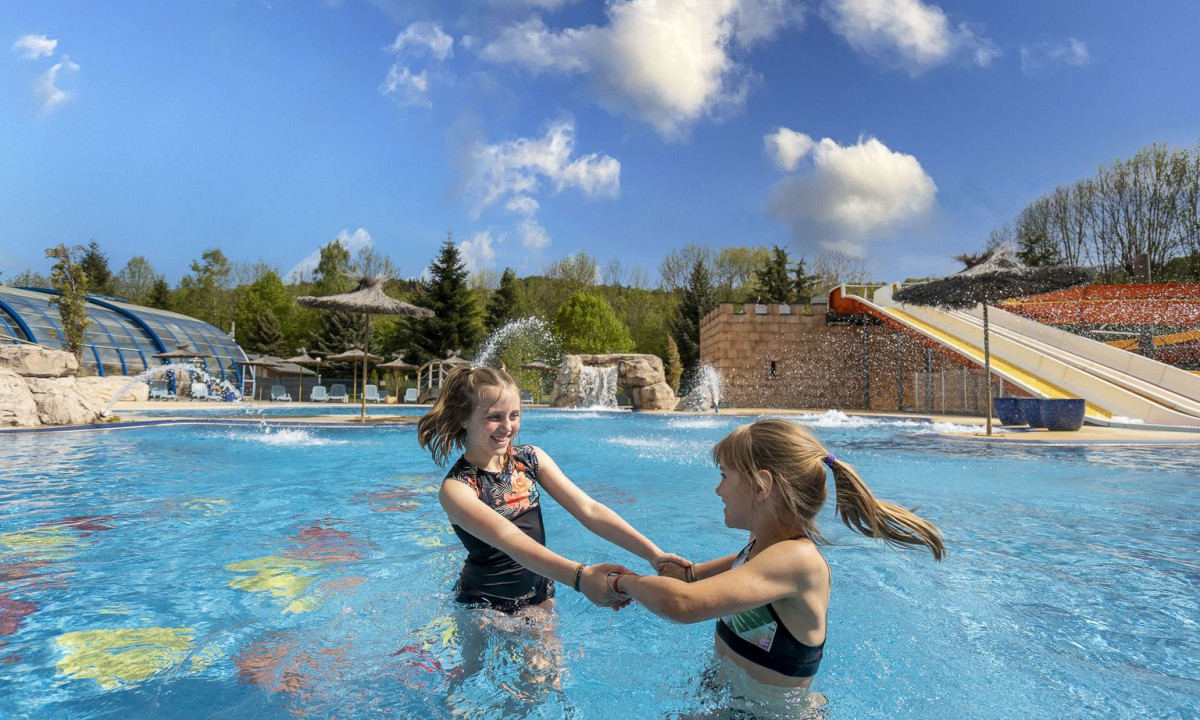 Two girls holding hands in a pool, splashing water, surrounded by slides, fountains, and trees at La Ribeyre