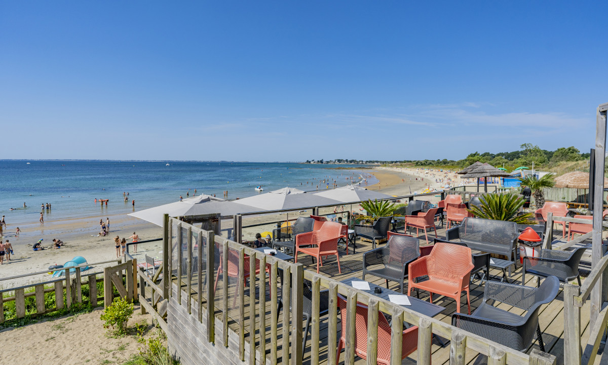 At De la Plage, a seaside terrace featuring vibrant chairs provides a view of individuals relishing the ocean and sandy beach beneath clear azure skies.