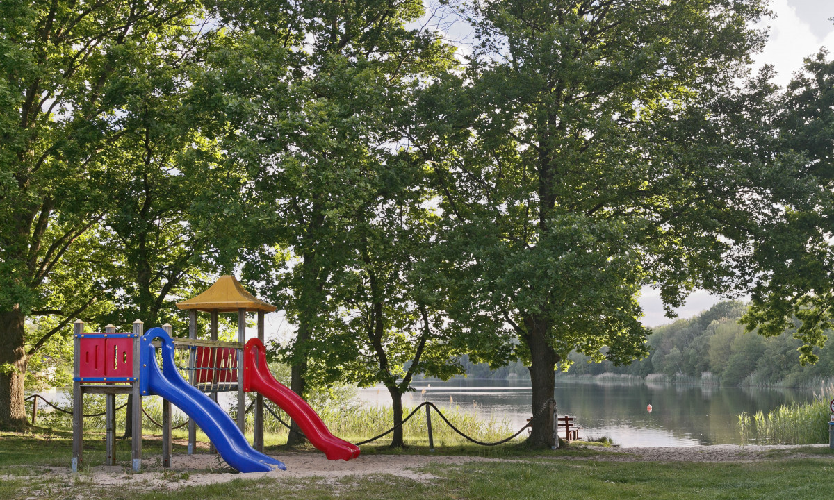 At KNAUS Campingpark Oyten, there is a play area featuring slides situated beneath trees next to a lake.
