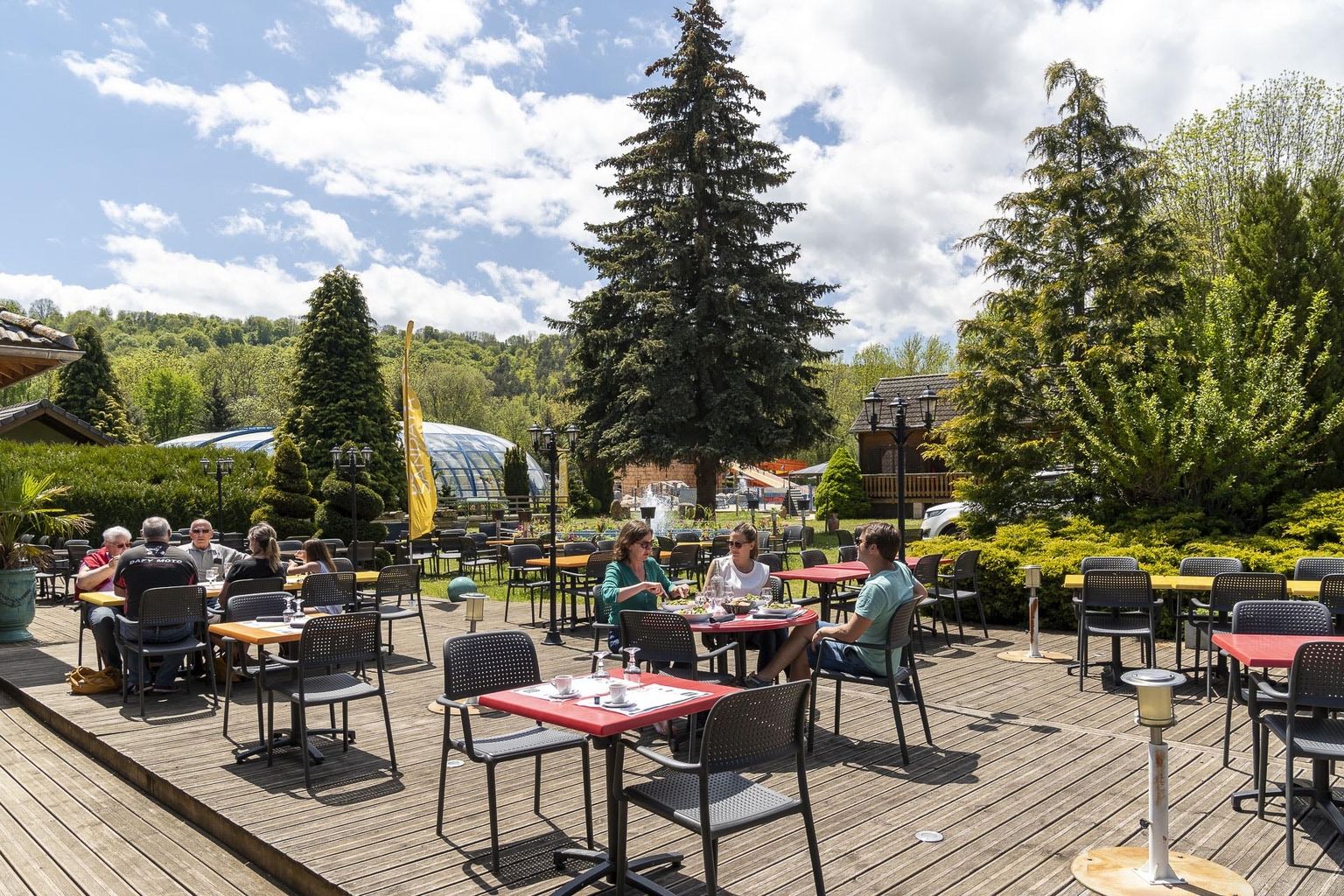 At La Ribeyre, individuals are enjoying a meal on an outdoor wooden terrace encircled by trees and various buildings.