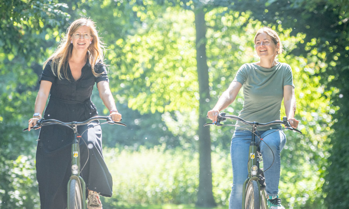 At Urban Gardens Gent, two individuals cycle, relishing a bright day along a tree-shaded trail.