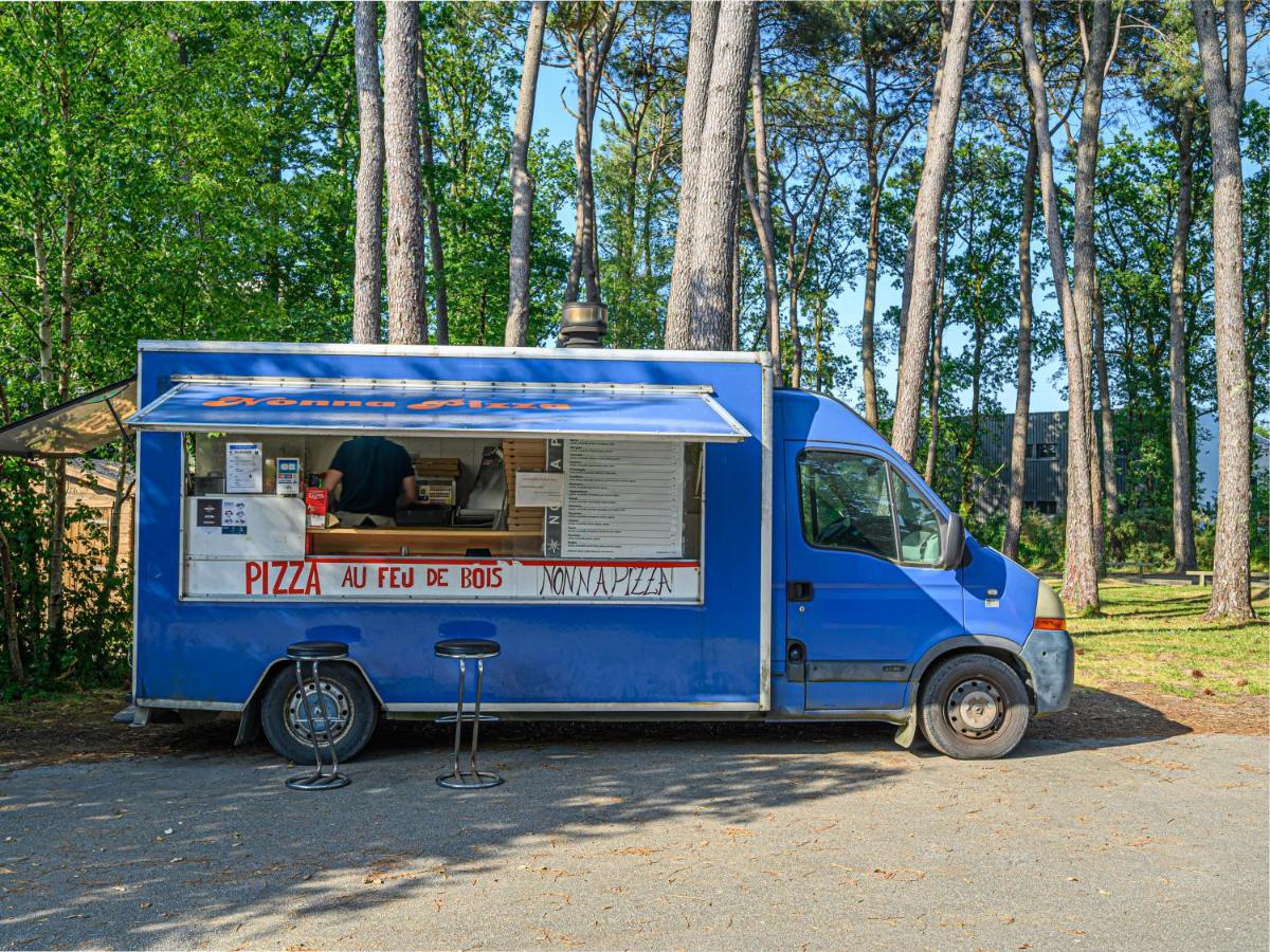 A blue food truck is selling wood-fired pizza, with barstools in front, surrounded by tall trees at Ker Goh Lenn