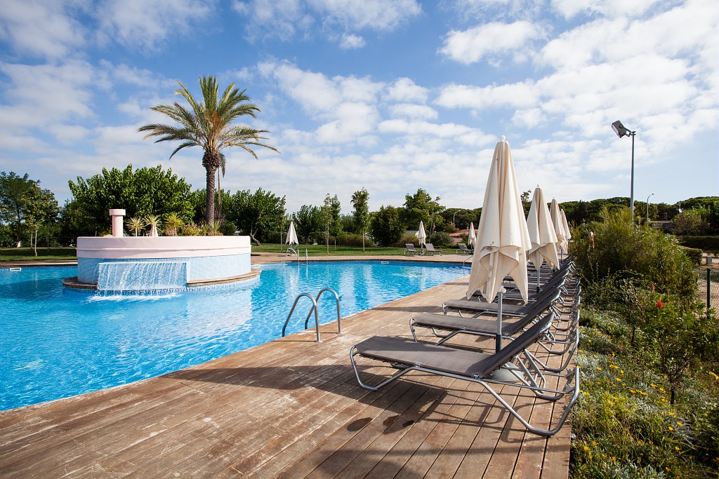 Pool with water fountain, surrounded by lounge chairs and umbrellas on wooden deck, lush green trees in background at El Pinar