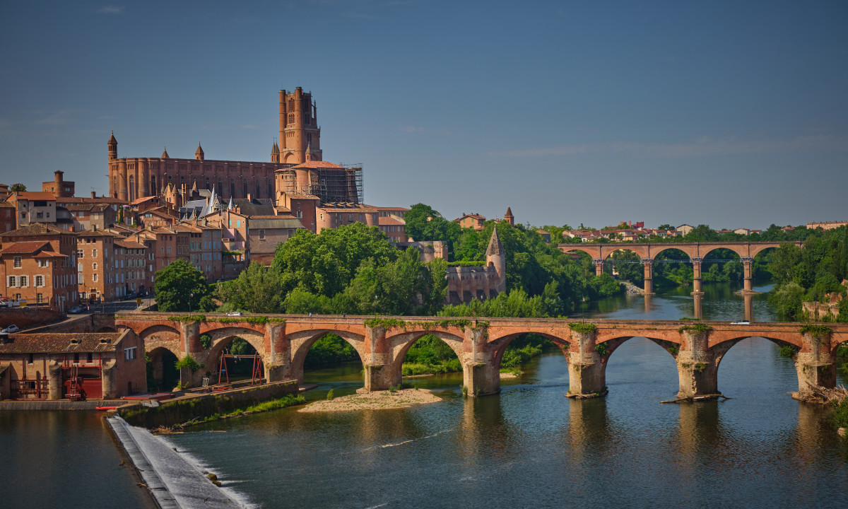 At Le Chêne Vert, a crimson brick cathedral looms over a historic town beside arched bridges spanning a river in a verdant, green setting.