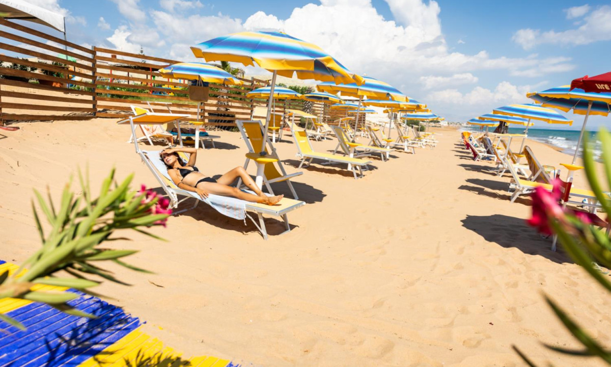 Woman sunbathing on a lounge chair under an umbrella on a sandy beach with rows of similar setups at Triscina Mare