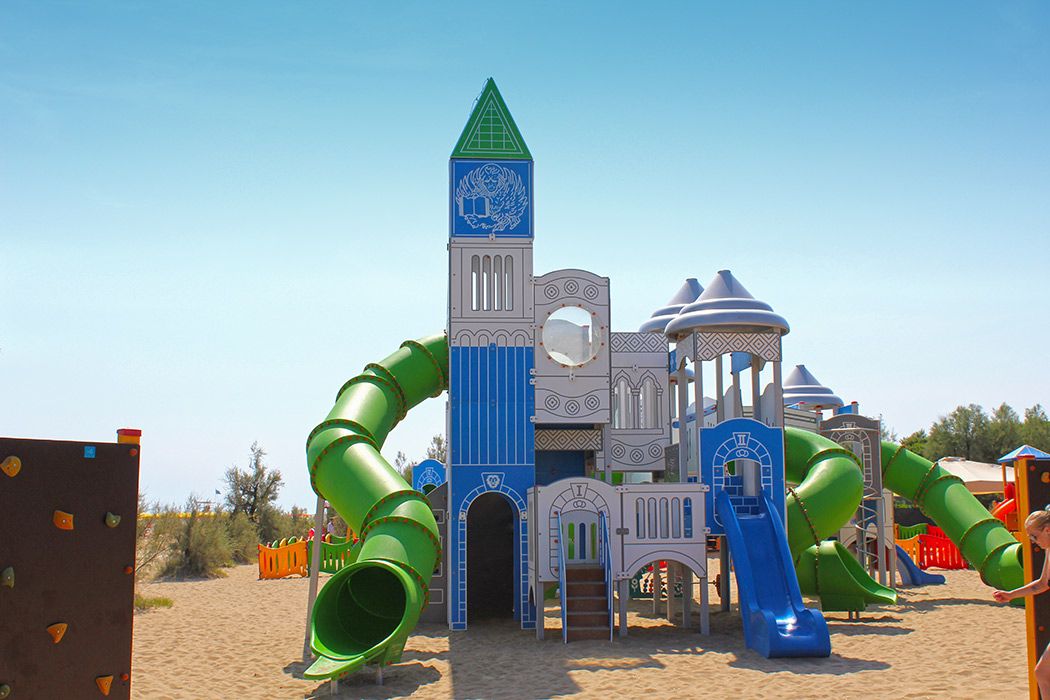 Castle-themed playground structure with slides and towers in a sandy play area, surrounded by trees and a climbing wall at Residence Village