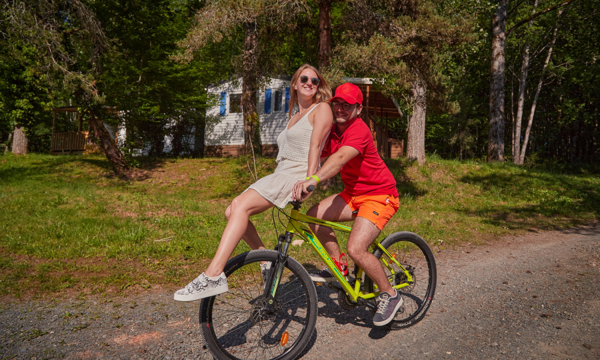 At Coucou, two individuals pedal a bicycle together along a dirt trail, encircled by trees and a cabin.