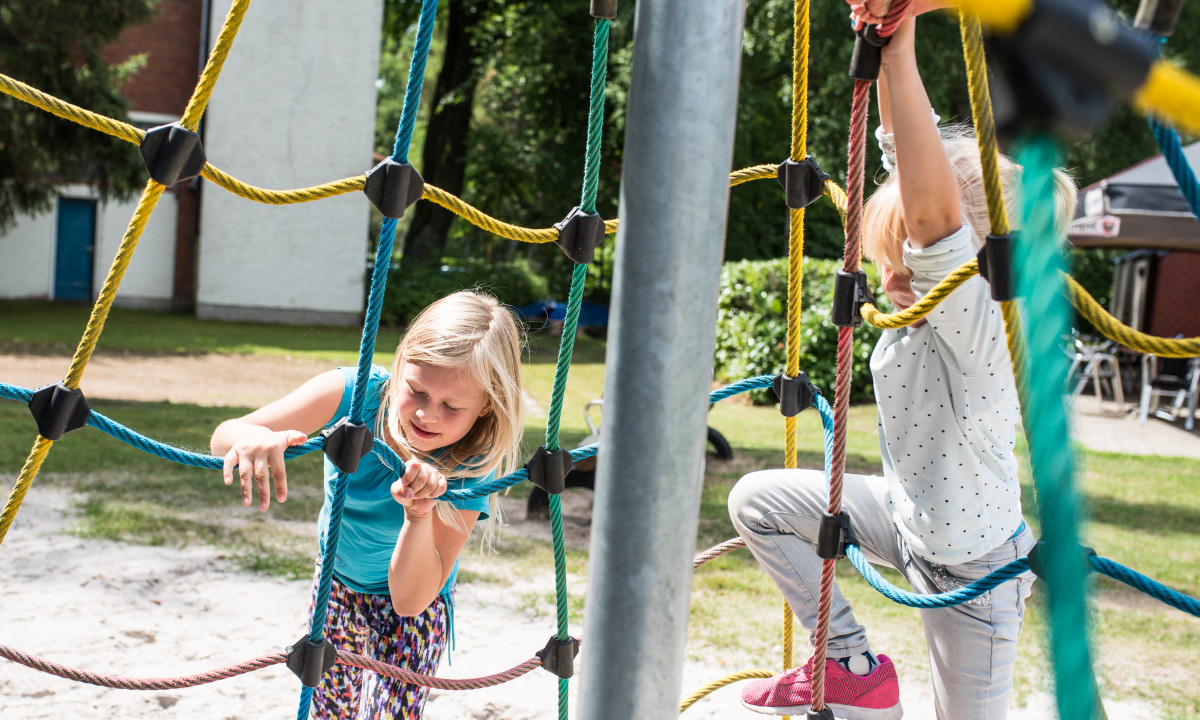 At het Veen, youngsters are scaling a vibrant rope net framework in a play area, with trees and structures in the backdrop.
