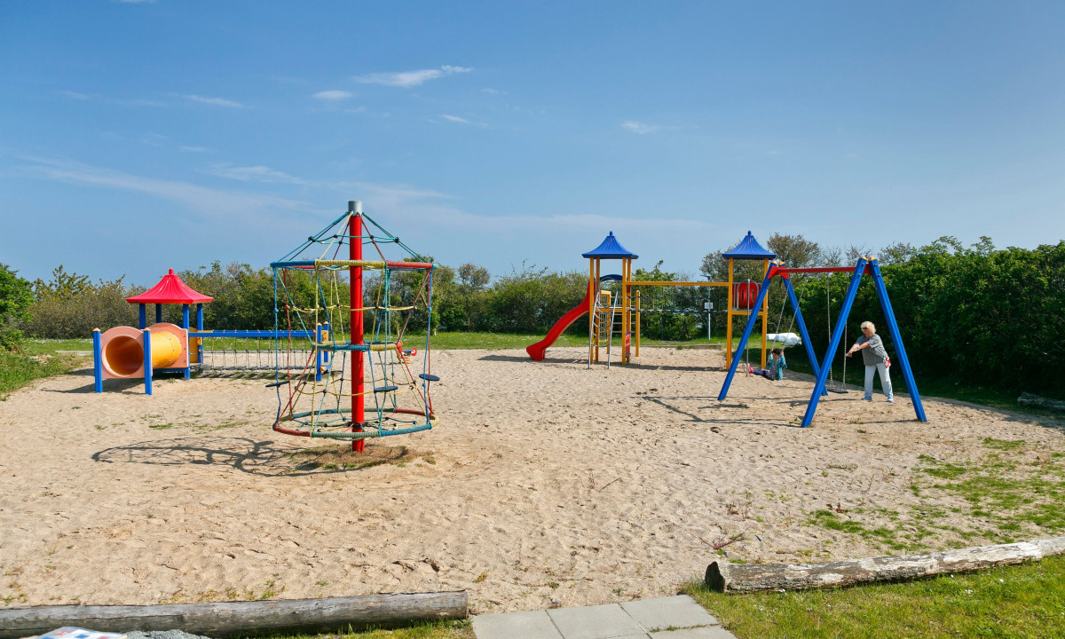 At KNAUS Campingpark Rügen, there is a children's play area featuring slides, swings, and climbing structures on a sandy ground, encircled by lush greenery.