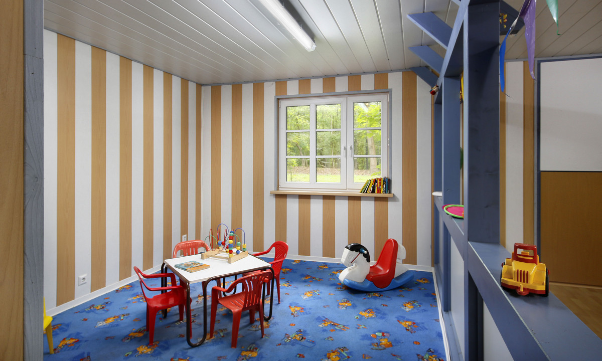 At KNAUS Campingpark Wingst, a children's table accompanied by red chairs is present, with toys strewn across a blue carpet in a playroom featuring striped walls. The window offers a view of lush greenery.