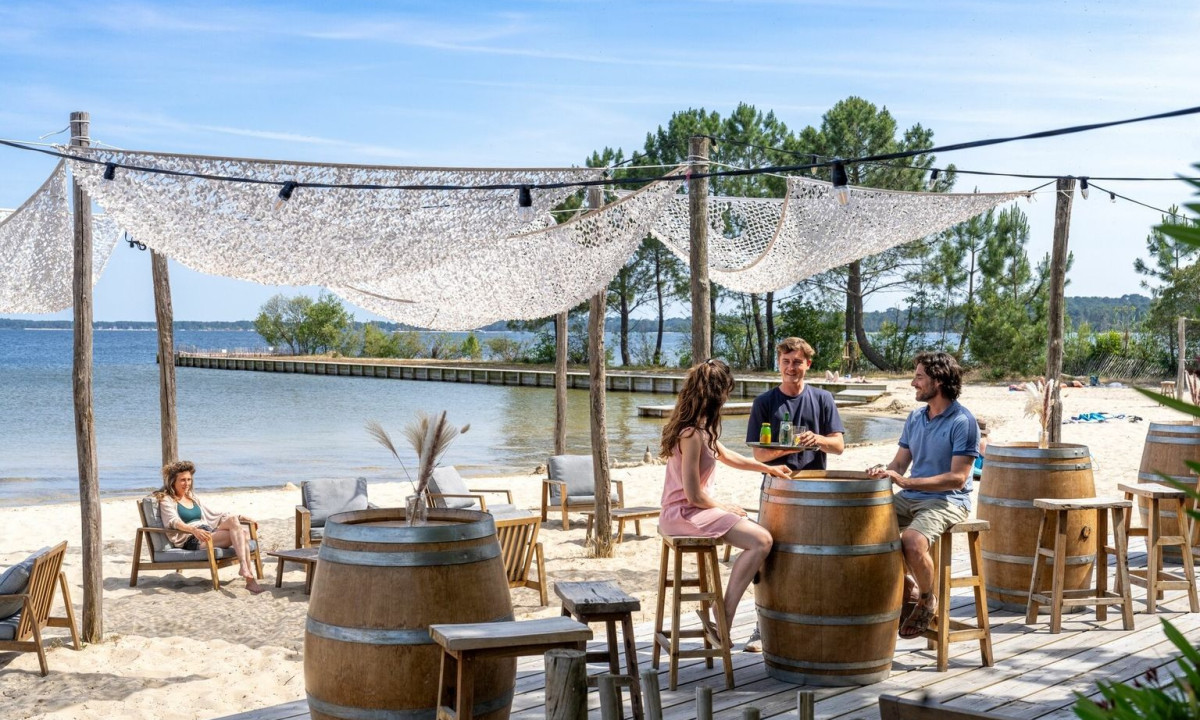 People sitting at a bar with barrel tables, enjoying drinks by a sandy lakeside with trees at Maguide