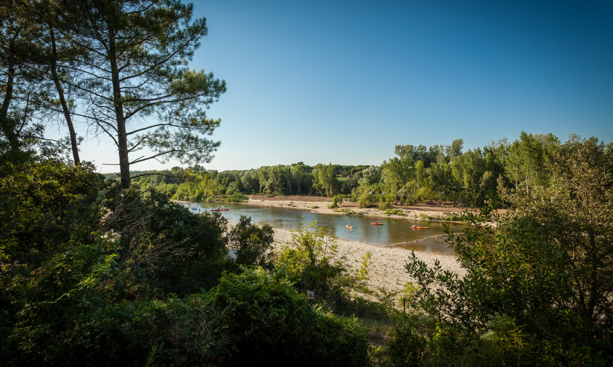 At La Sousta, kayaks drift on a tranquil river encircled by vibrant green trees and a clear azure sky.