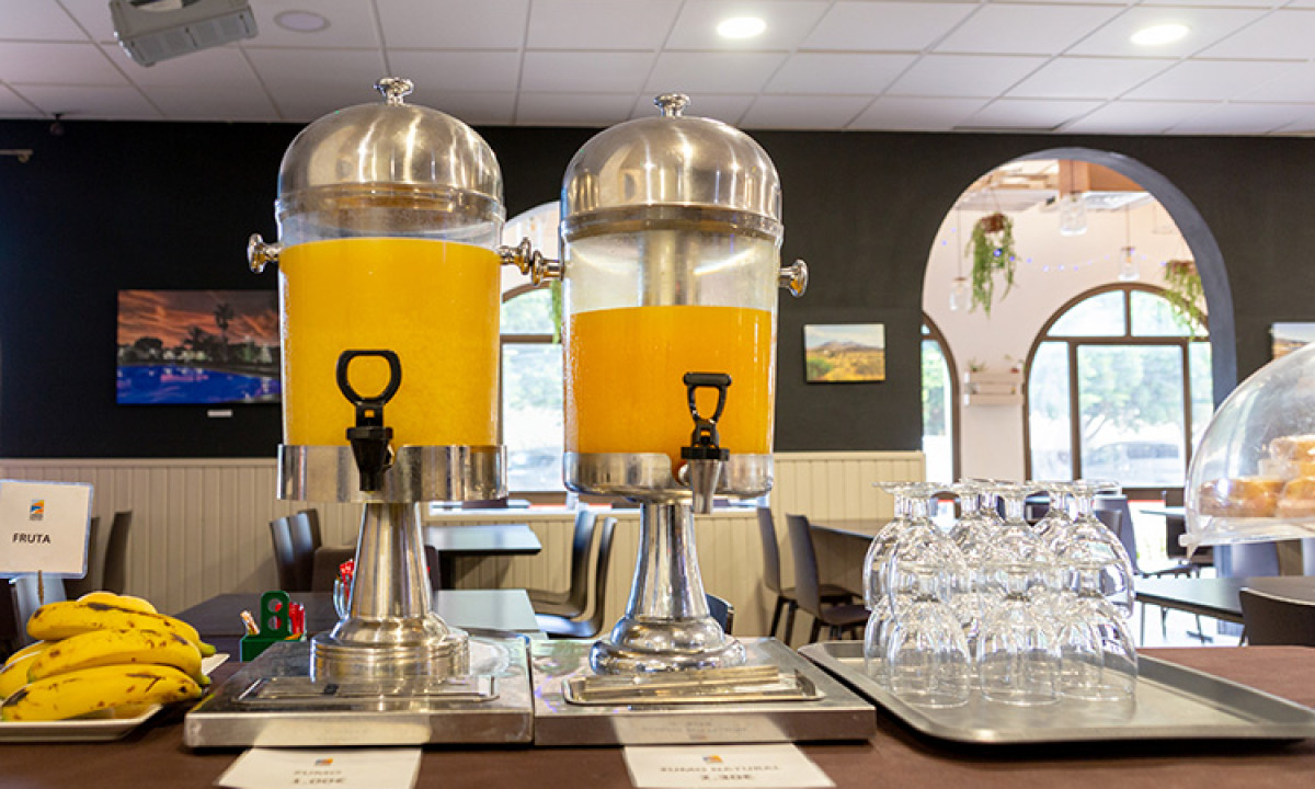 At Camping Los Escullos, two sizable juice containers brimming with orange liquid are placed on a buffet table, accompanied by glasses and bananas.