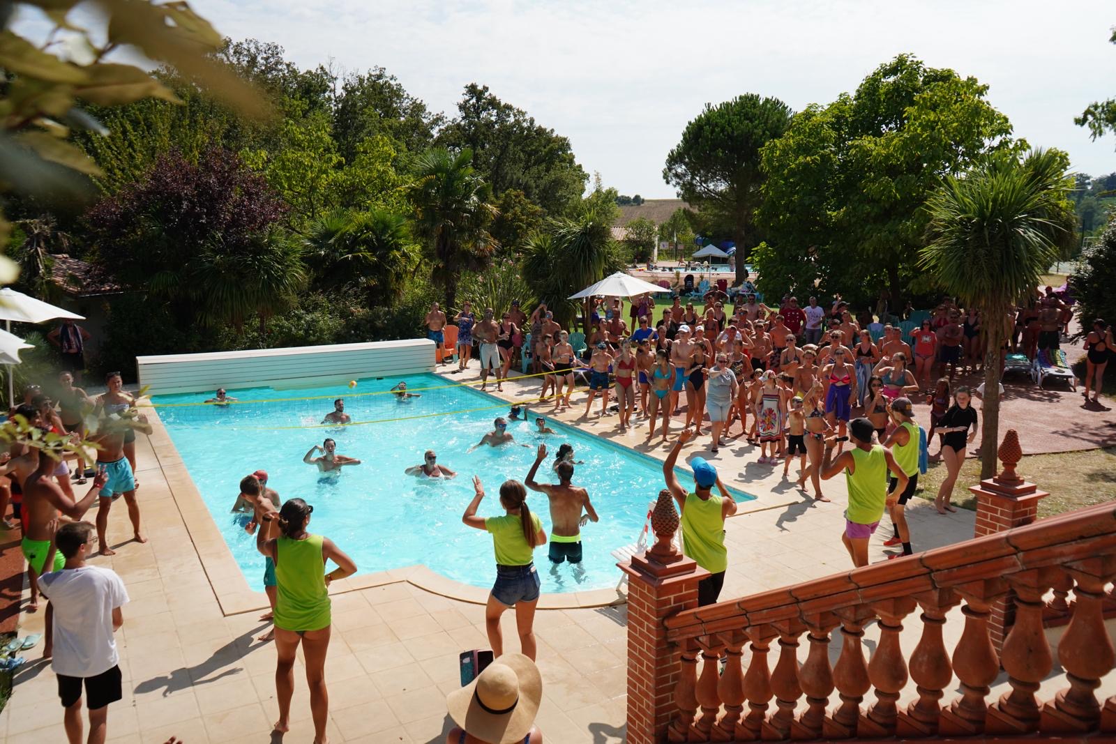 At Domaine du Koukano, individuals in vibrant swim attire are engaging in activities in a swimming pool, while an audience observes from beneath trees and umbrellas.