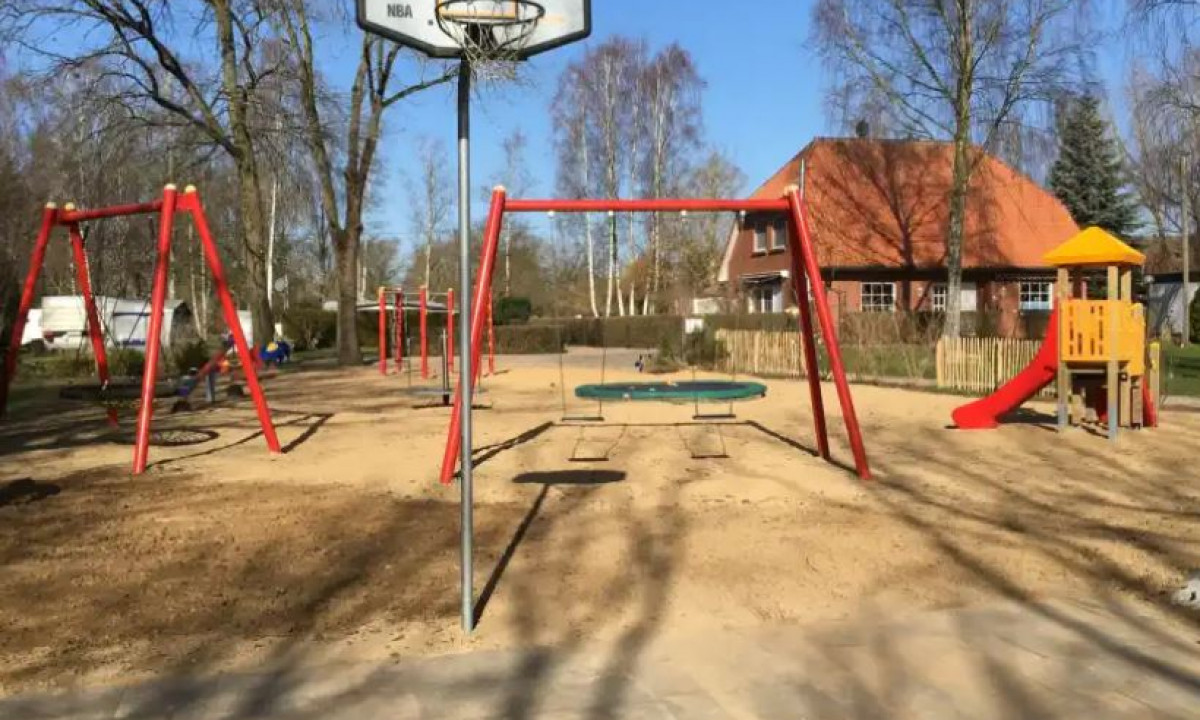 At Elbeling, there is a play area that includes swings, a slide, and a basketball hoop, located in a sandy space next to trees and residences.