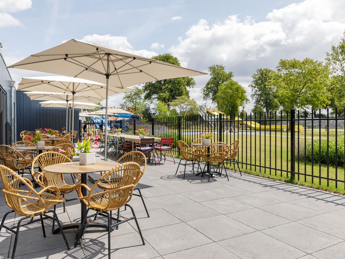 At De Twee Bruggen, wicker seating and tables adorned with blossoms are placed beneath large canopies on a terrace adjacent to a fenced grassy space.