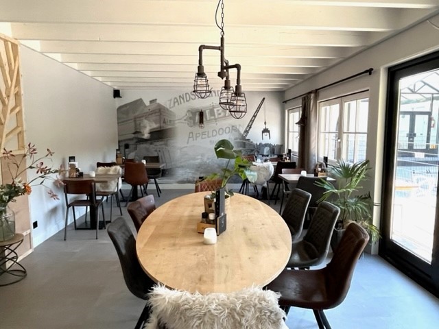 At de Vinkenkamp, dining tables accompanied by chairs and ornamental pieces are arranged in a contemporary, industrial-themed space featuring expansive windows and a mural.