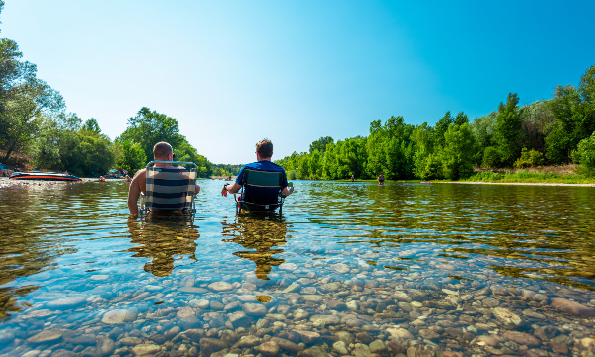 At La Sousta, two individuals are seated on chairs in shallow water, encircled by trees and a clear blue sky.