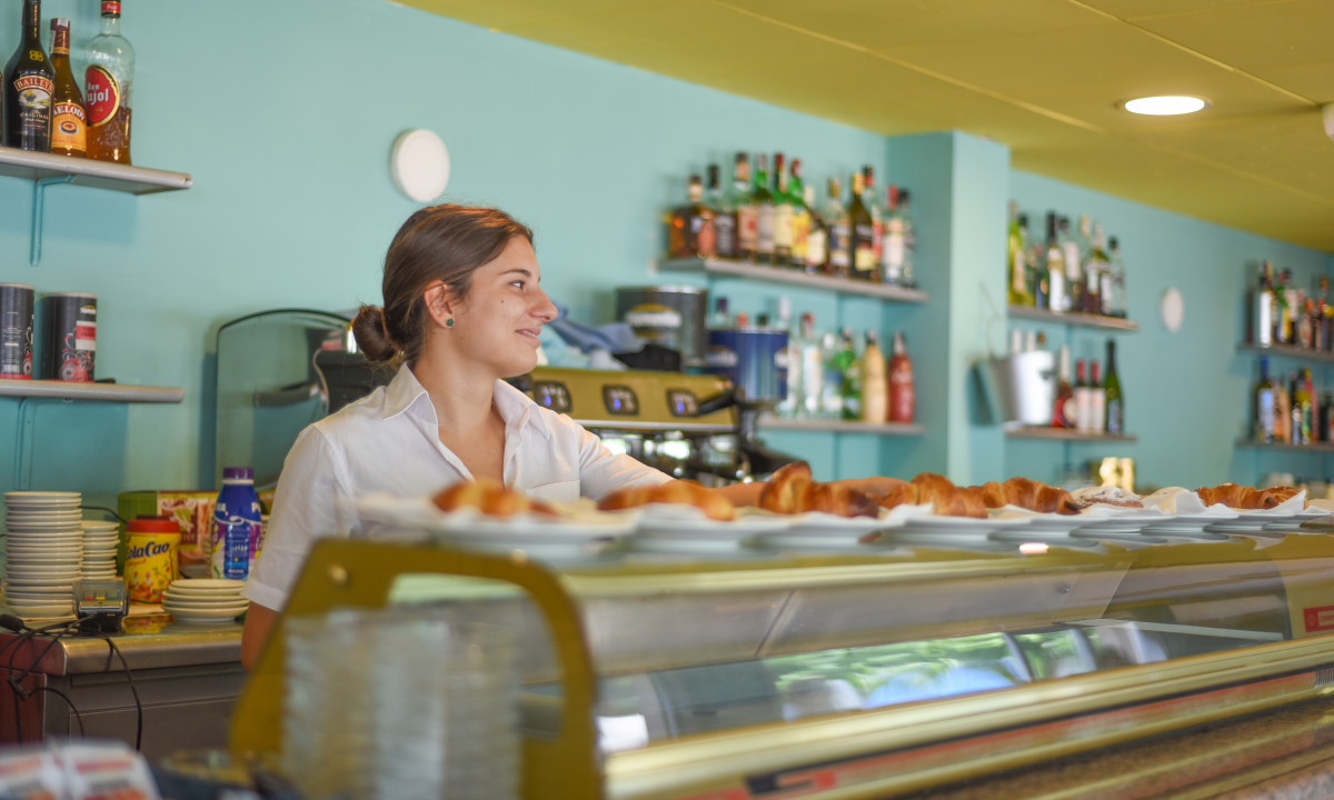 Barista smiling while serving pastries at a brightly lit, well-stocked café counter at Resort Els Pins