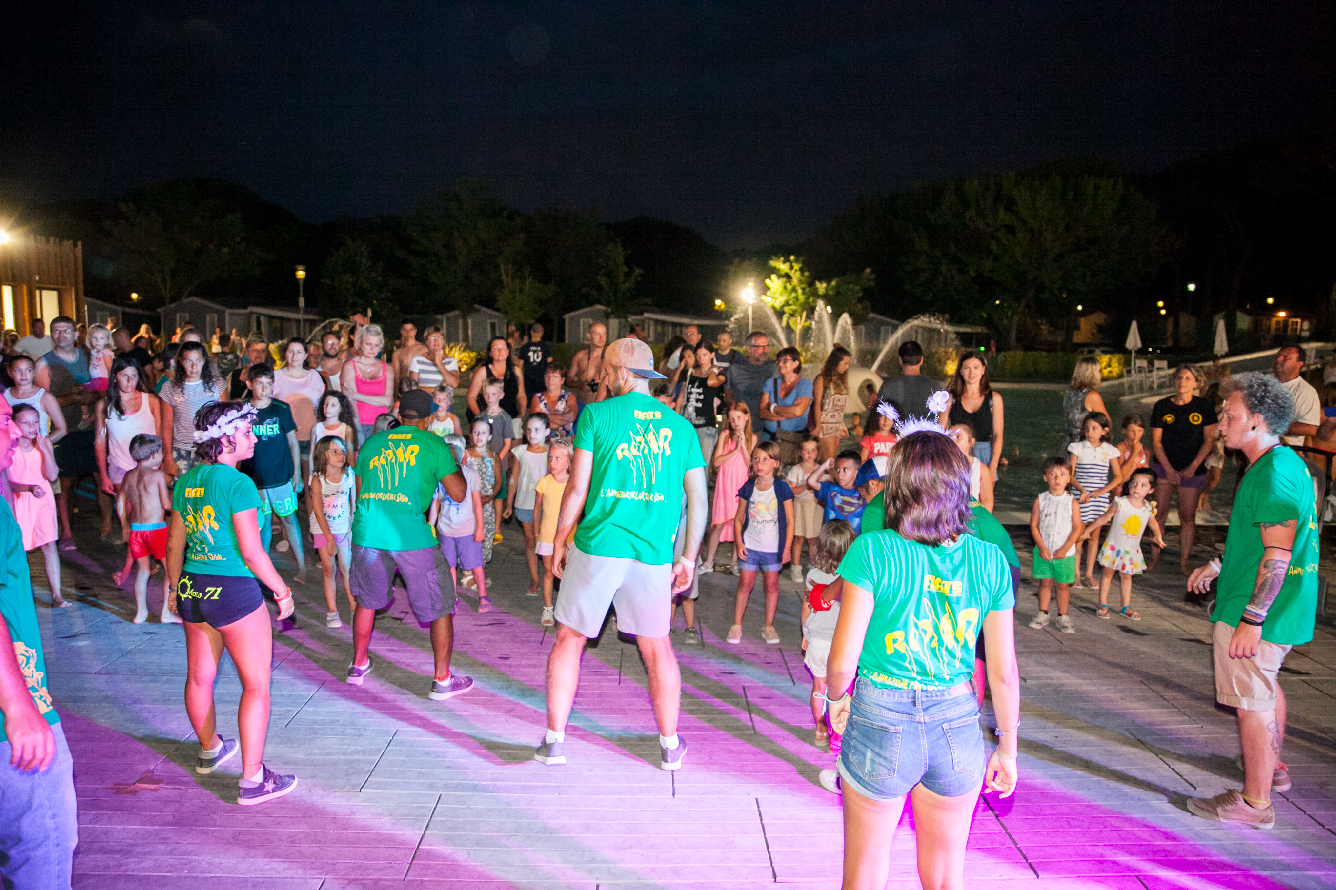 People dance in brightly lit group activity, joined by children and adults, nocturnal outdoor gathering at Pineta Sul Mare Camping Village