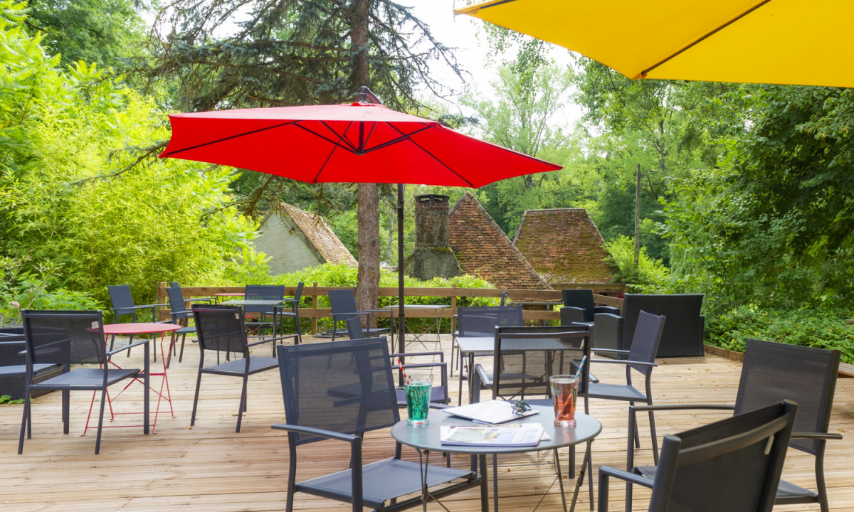 At Coucou, chairs and tables beneath vibrant umbrellas are situated on a wooden terrace, encircled by abundant foliage and charming rooftops.