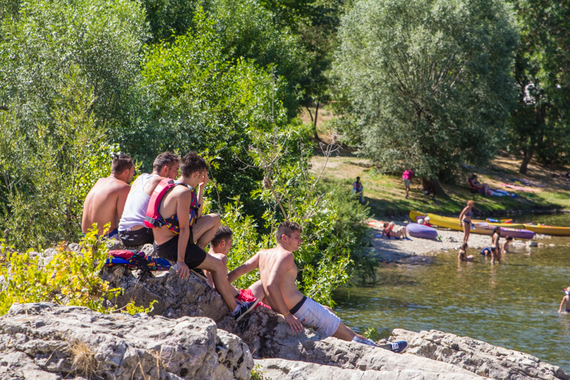 Group of people sitting on rocks, observing swimmers and kayaks by a riverside, surrounded by trees at du Domaine d'Anglas