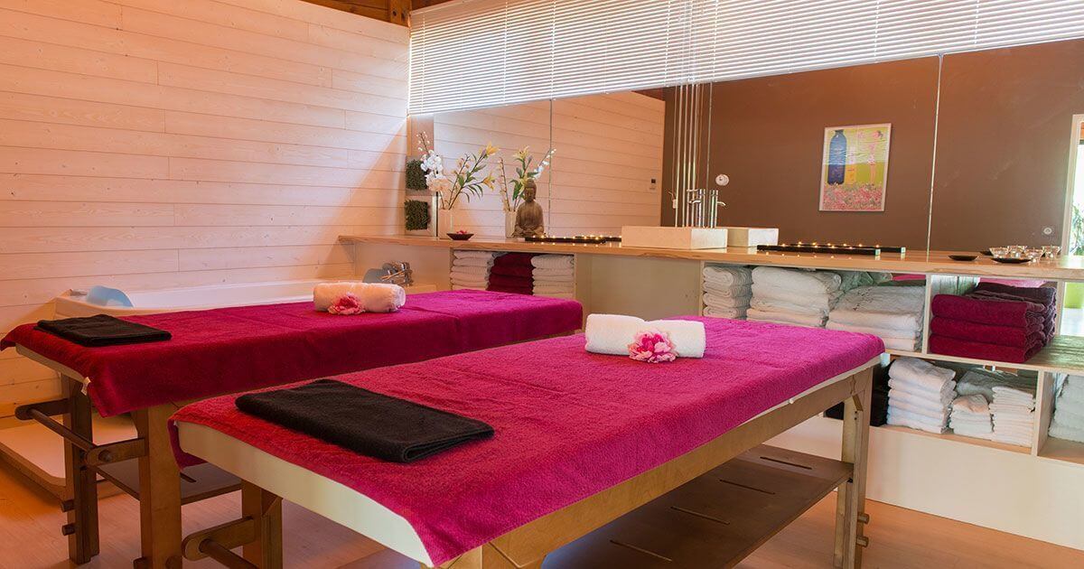At Le Ranc Davaine, two massage tables adorned with towels and blossoms are encircled by shelves filled with neatly arranged linens in a spa.