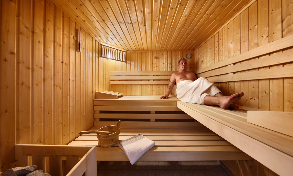 At KNAUS Campingpark Rügen, an individual unwinds on a timber bench in a sauna, with a towel draped around them, encircled by wooden walls and ceilings.