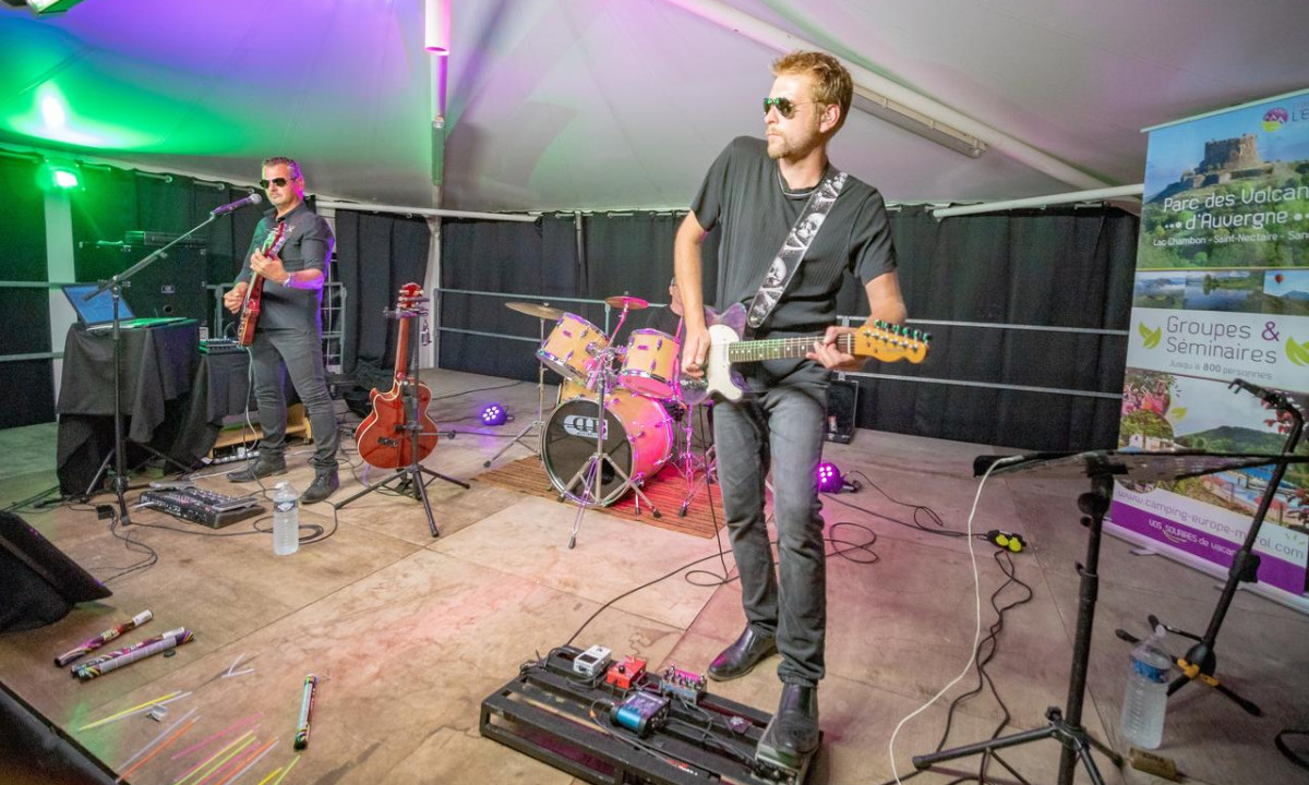 Two musicians play guitars on an illuminated stage with drums in a large tent at Paradis l'Europe