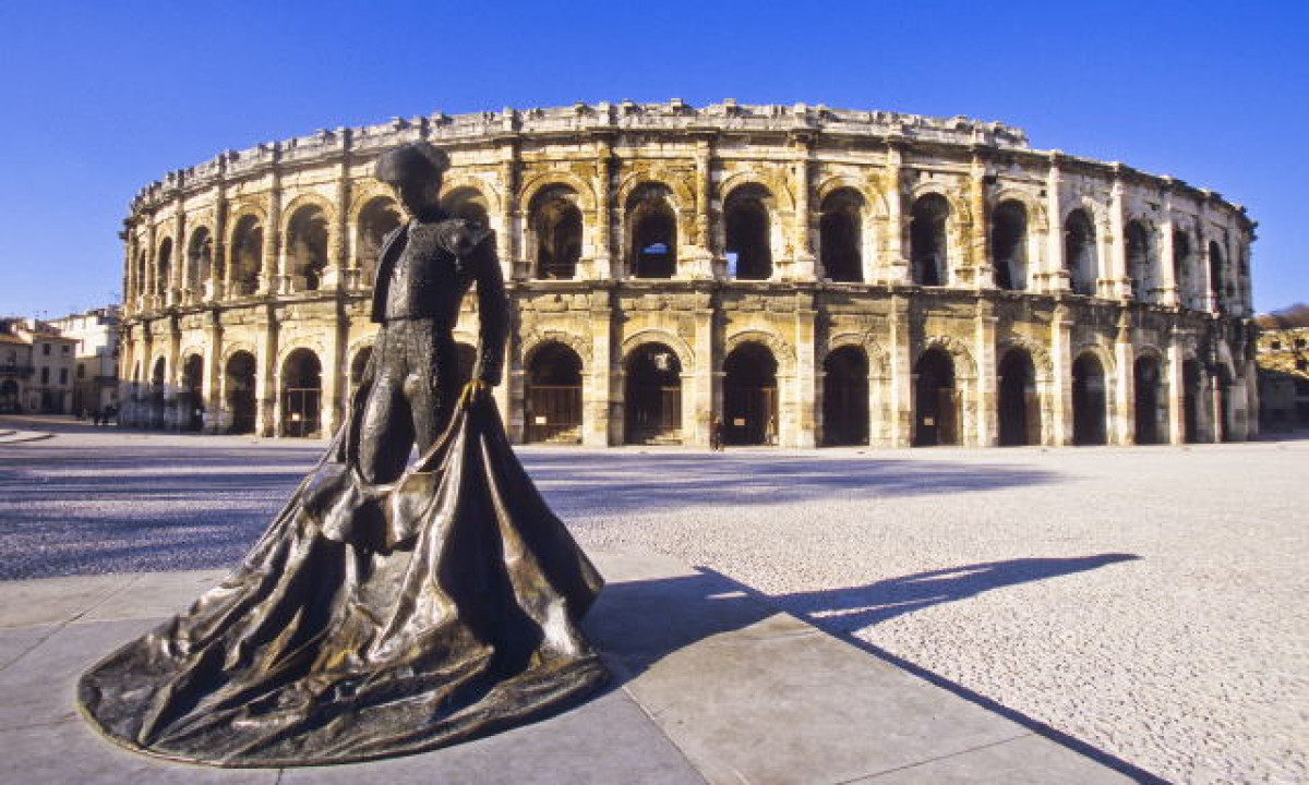 At La Clémentine, a bronze bullfighter sculpture stands in front of an ancient Roman arena beneath a clear azure sky.