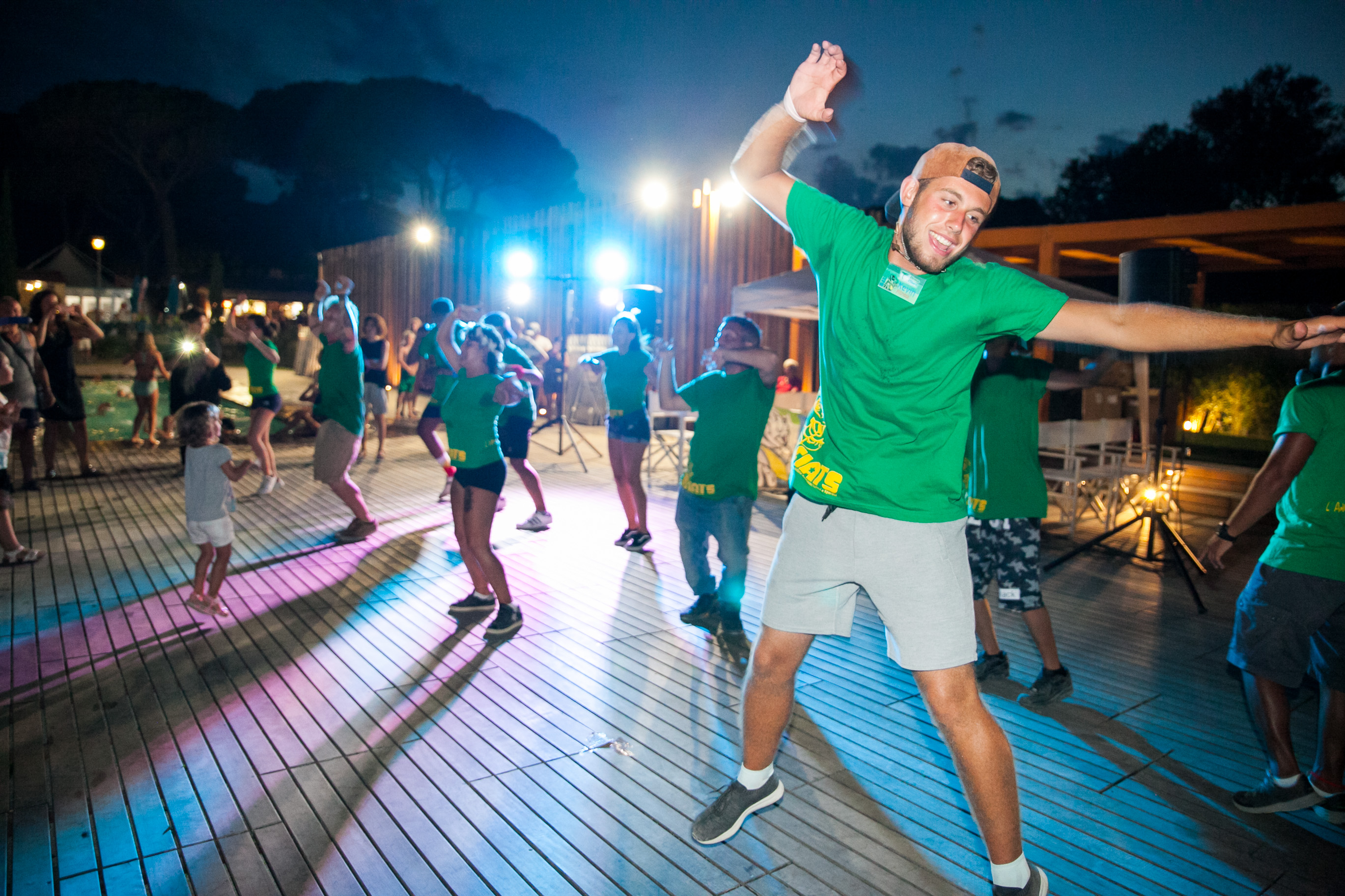 People dancing energetically under colorful lights on a wooden deck surrounded by trees and onlookers at Pineta Sul Mare Camping Village