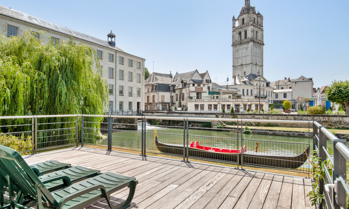 Deck chairs on a wooden terrace, overlooking a river with a gondola, flanked by historic buildings and a tall tower at Le Moulin des Cordeliers