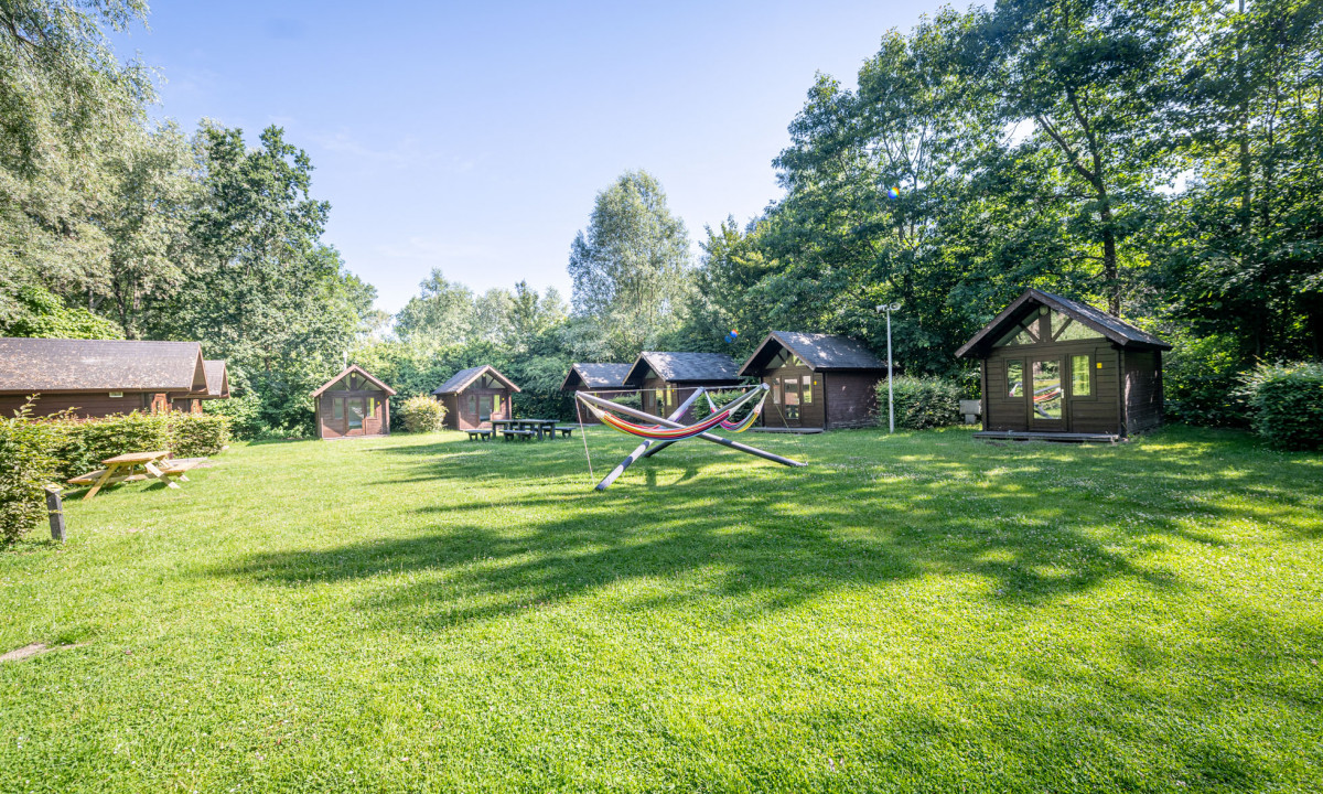 At Urban Gardens Gent, a hammock is positioned on a grassy lawn surrounded by a number of small wooden cabins amidst verdant trees.