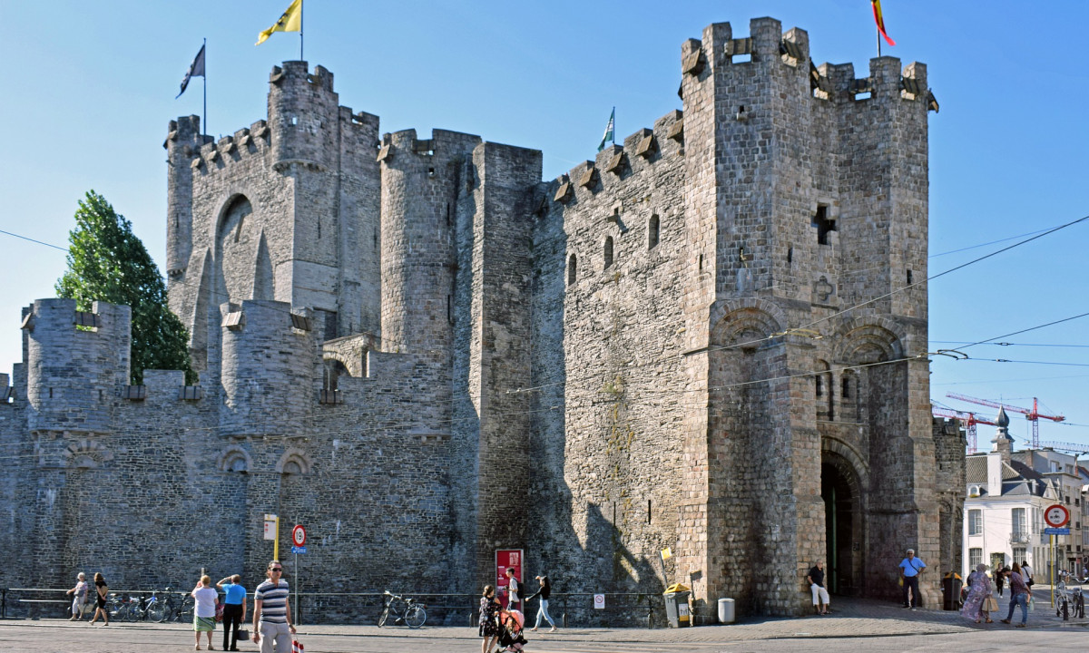 At Urban Gardens Gent, a stone fortress stands steadfast with banners waving, encircled by leisurely walkers and cyclists in a bright city plaza.